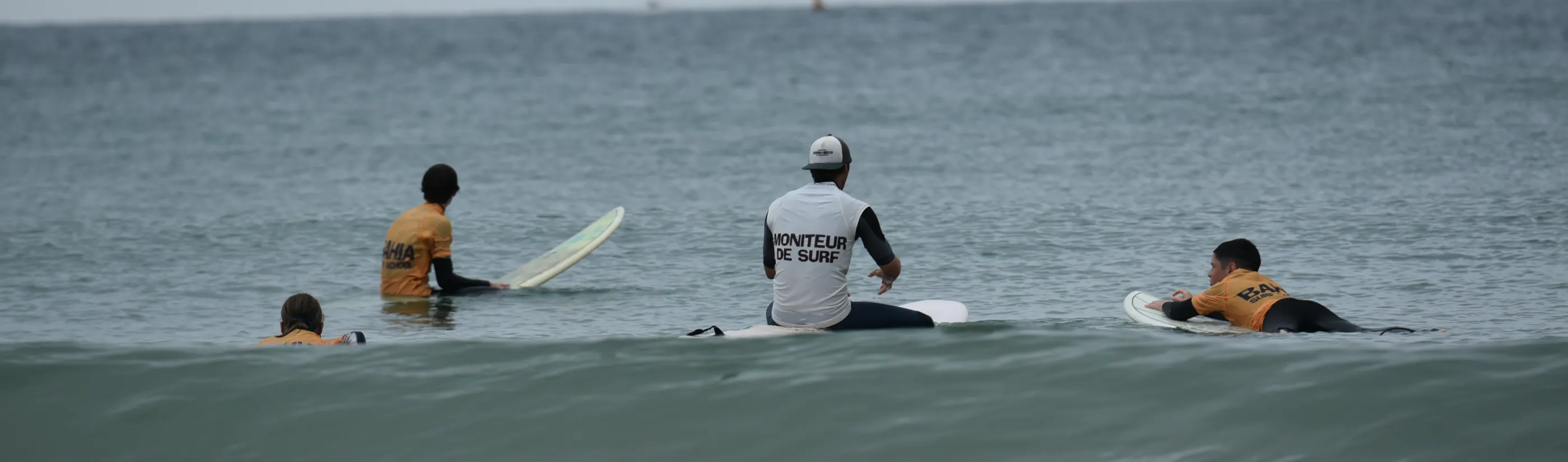 Le moniteur assis sur sa planche au large, dossard « moniteur de surf » dans le dos, trois élèves en lycra Bahia autour de lui.