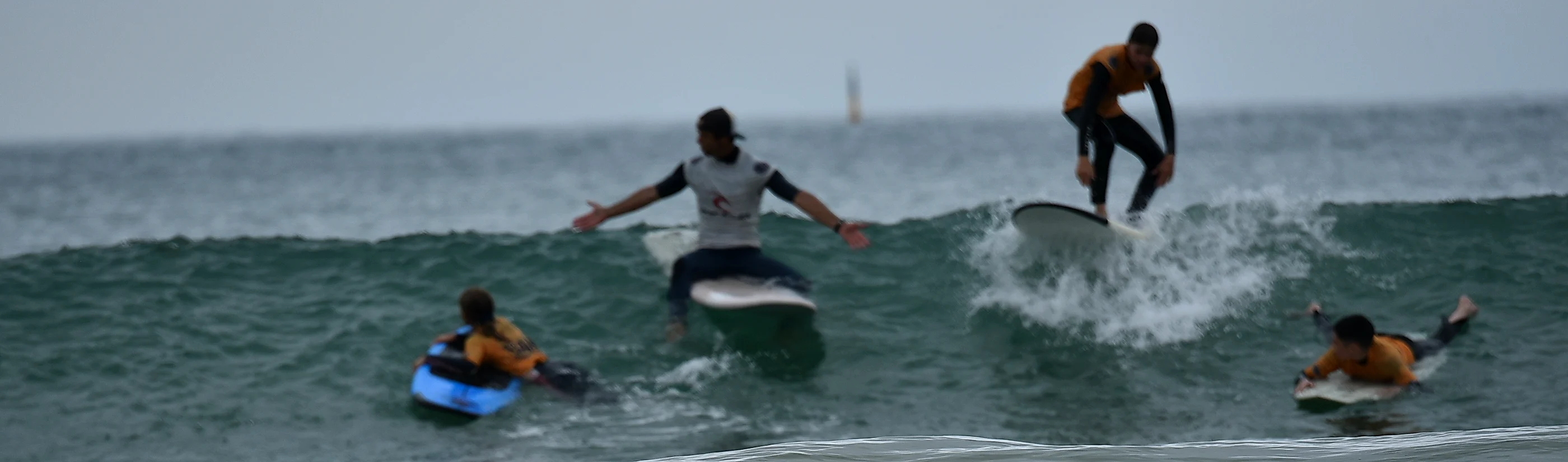 Une session au large : le moniteur en équilibre sur sa planche, un élève debout sur la vague à côté, deux autres allongés qui rament.
