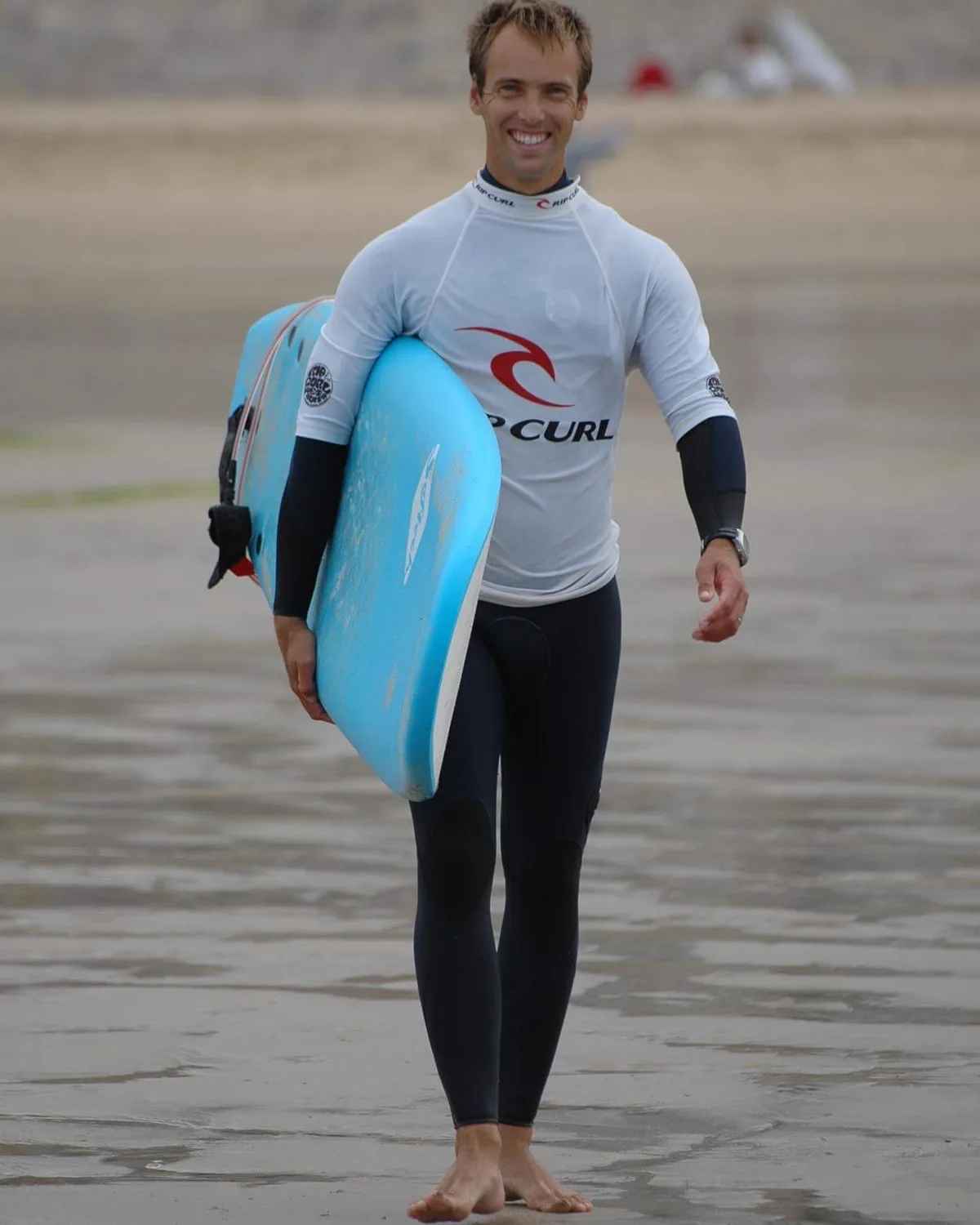 Simon Praud remonte la plage en combinaison, sa planche sous le bras, souriant.