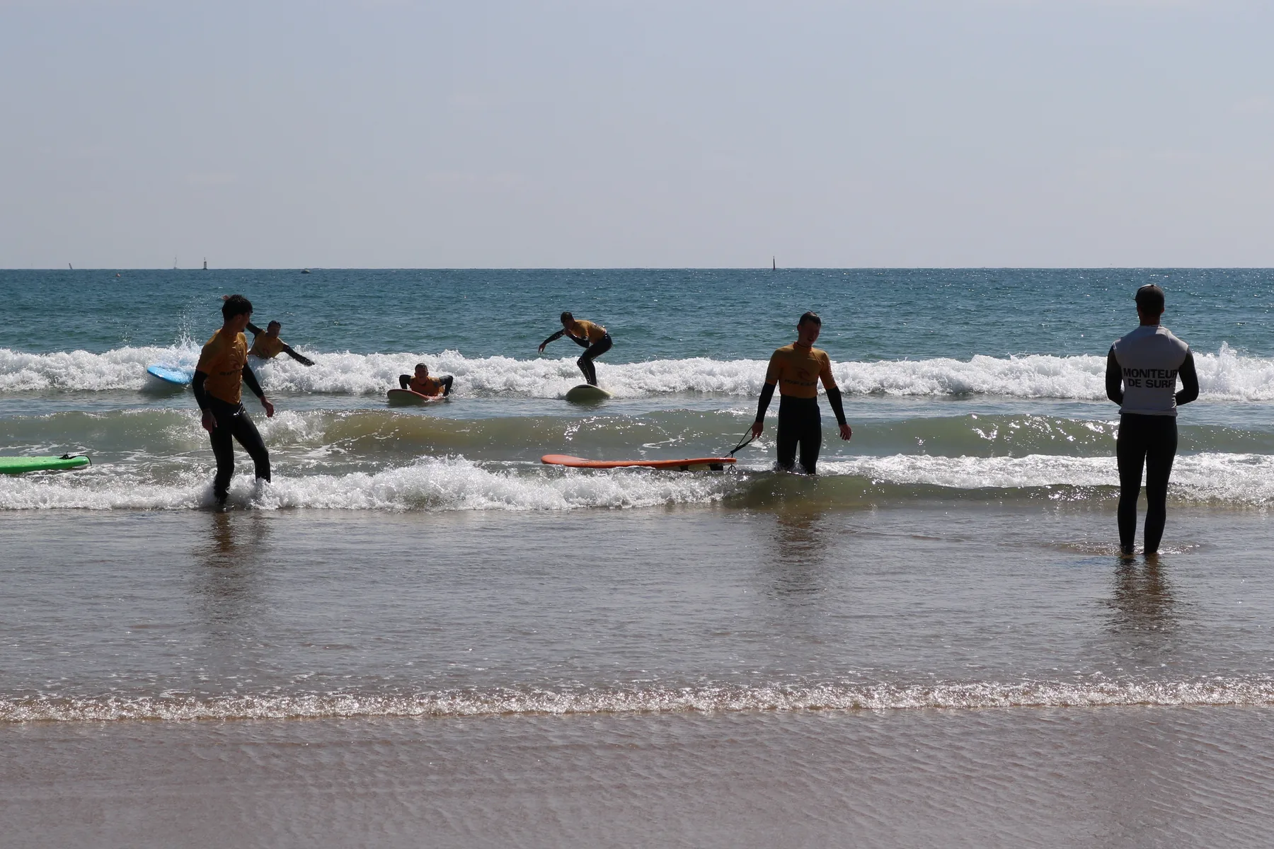 Les élèves repartent dans la mousse avec leurs planches, le moniteur debout dans l’eau les regarde.