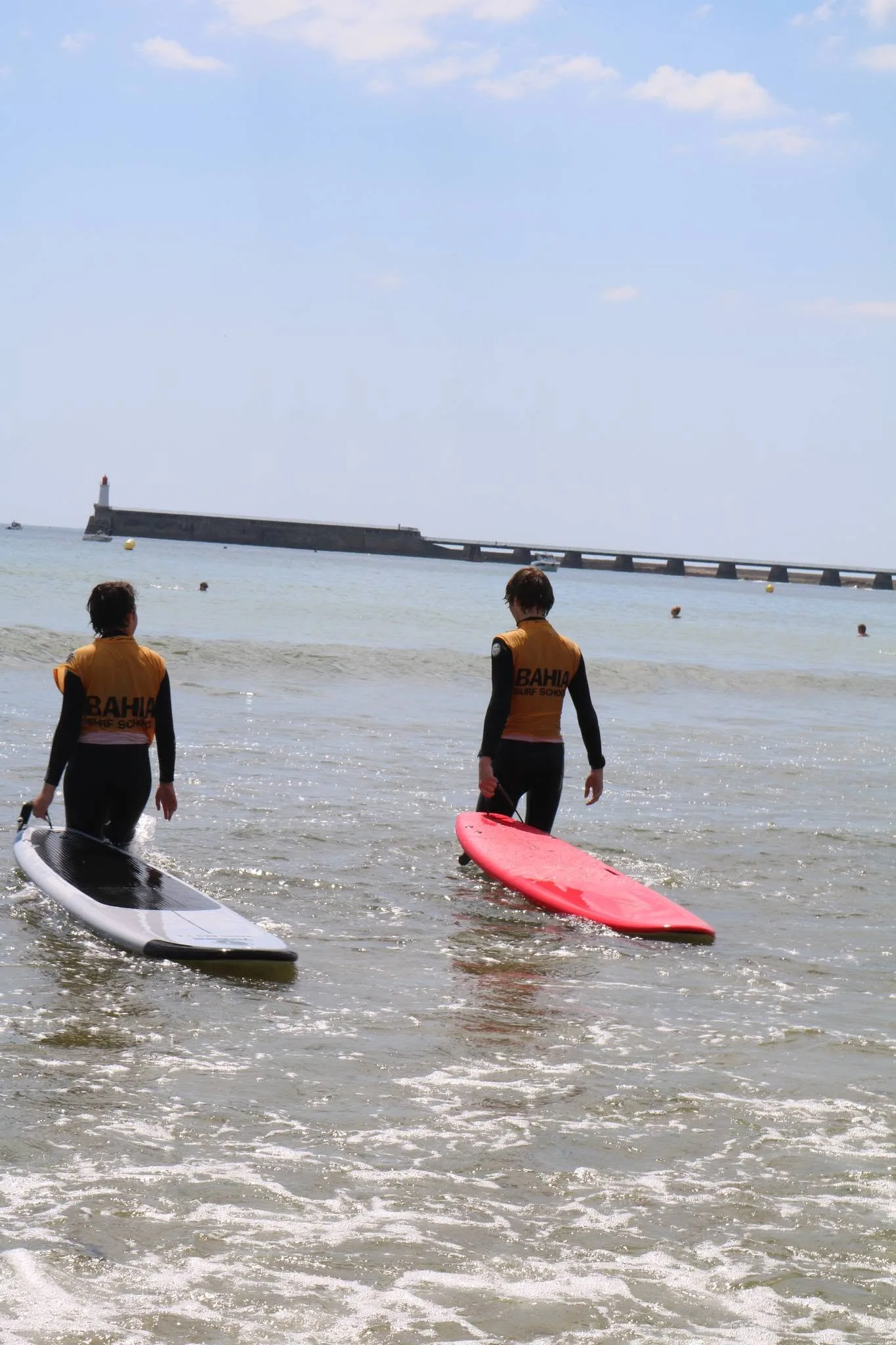 Deux élèves entrent dans l’eau, planche sous le bras, la jetée au fond.