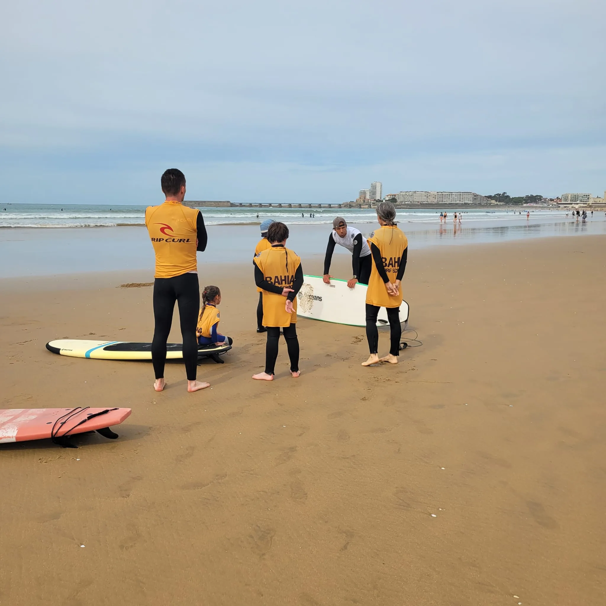 Le moniteur avec un enfant et une élève qui porte sa planche, au bord de l’eau.