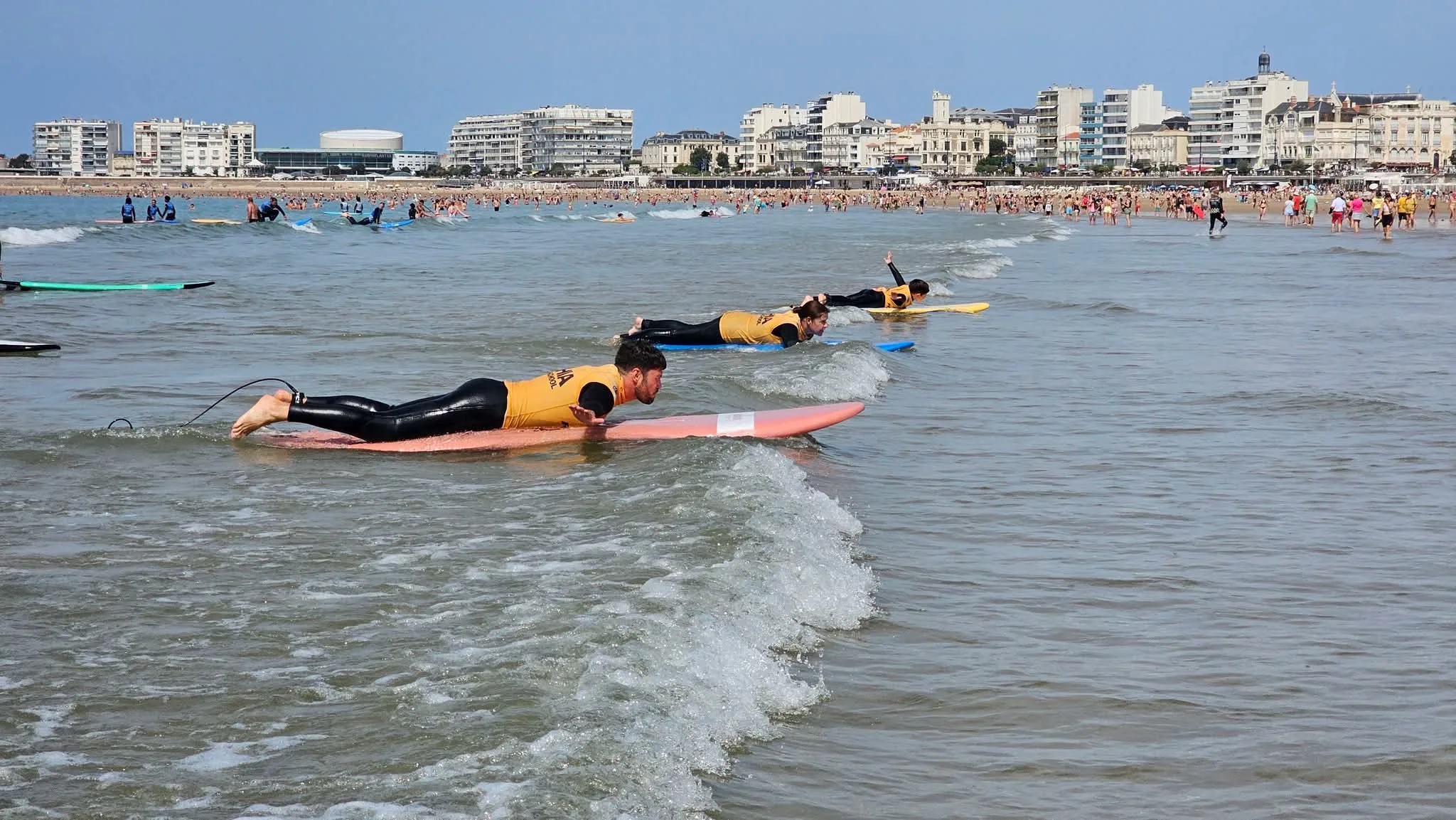 Trois élèves à plat ventre rament dans la mousse, le front de mer derrière.
