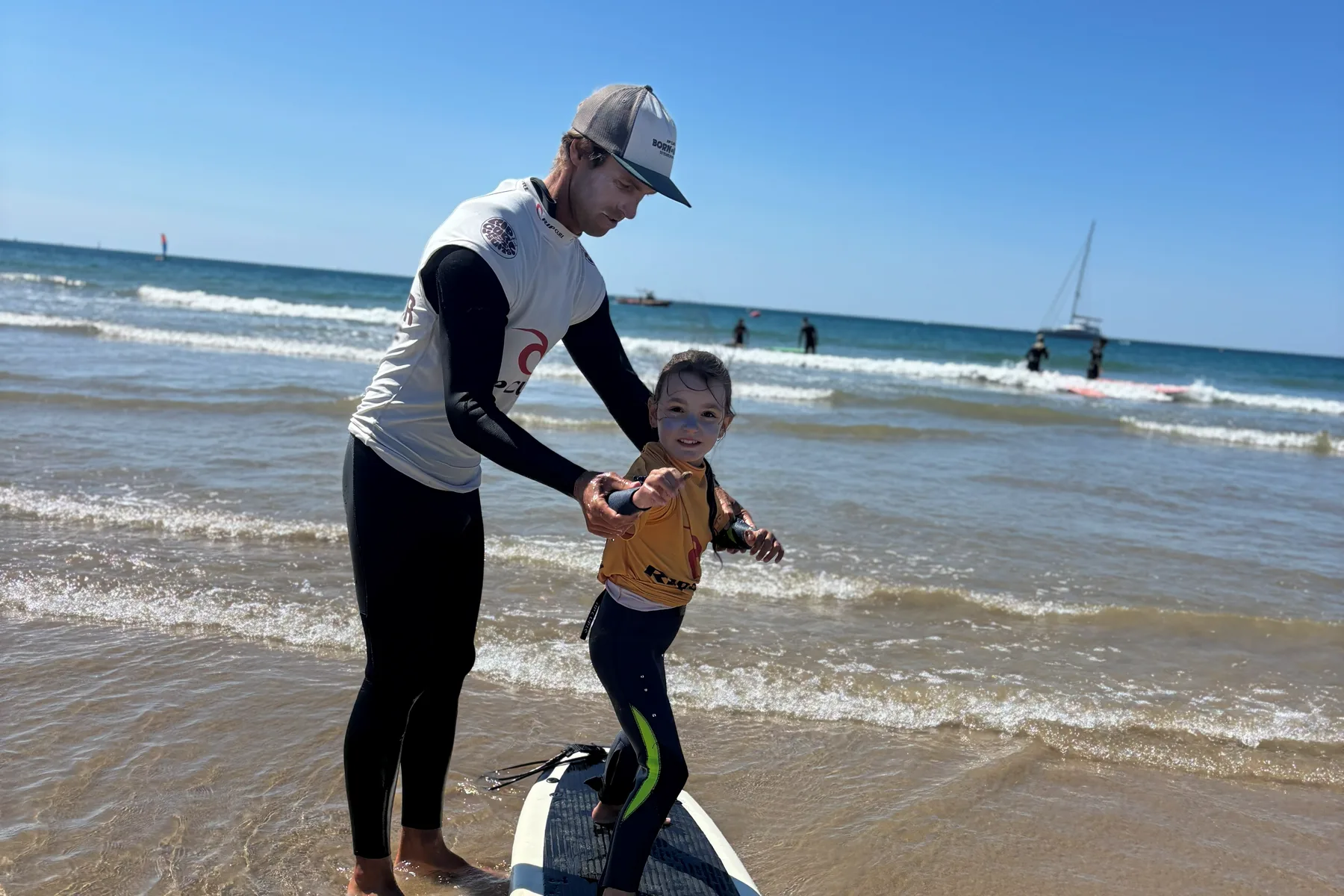 Simon tient les mains d’un enfant debout sur une planche, au bord de l’eau.